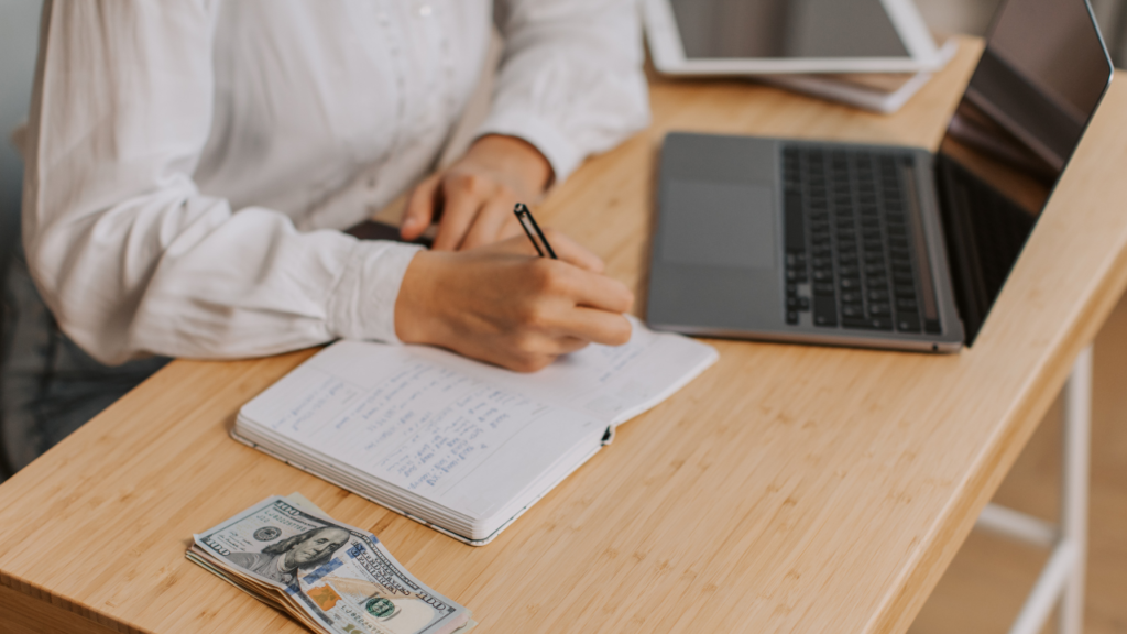 person writing in notebook with computer and cash on desk