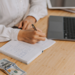 person writing in notebook with money and computer on desk