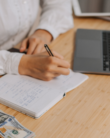 person writing in notebook with money and computer on desk